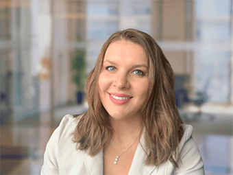 Shelby has wavy light brown hair with blonde highlights. It falls around her shoulders loosely. She wears a white coat. She has a silver necklace. She is smiling.