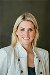 Kari has hazel green eyes. She has blonde hair parted in the middle. She is wearing a white blazer jacket with bronze buttons and a black blouse underneath.