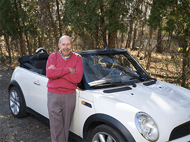 Bob stands in front of a white convertible looking happy. He wears gray slacks and a red sweater.