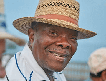 Emerson smiles. He wears a straw woven hat. He has on a white and blie striped shirt. The sky is blue behind him.