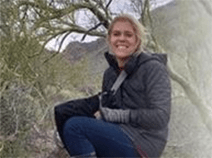 Ashly sits on a rock in a forest. She has on boots, jeans, and a black coat. She has blonde hair and she is smiling.