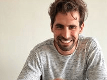 Dr Isaac Cohen smiles. He sits at a table with a bowl of soup and a glass of wine. He has brown hair and a beard. He wears a gray long sleeve shirt.