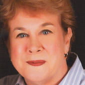 A woman smiles. She has short red hair, blue eyes, and wears a striped collared shirt and bright red lipstick.