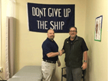 Two men stand in a physical therapy office. One man wears jeans and a black shirt, while the other wears khakis and a black shirt. There is a chiropractic table beside them.