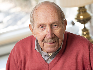 Bob stands in front of a white convertible. He looks happy. He wears a red sweater and gray slacks.