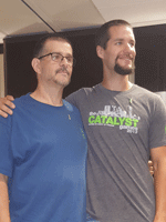 Two men stand side by side. The man on the left wears a blue shirt, and the man on the right wears a gray shirt. The man in gray puts his arm around the other man's shoulder in a friendly gesture.