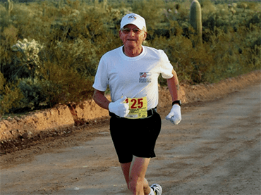 A man with gray hair and rounded glasses. He wears a black shirt. He is smiling and looks happy.